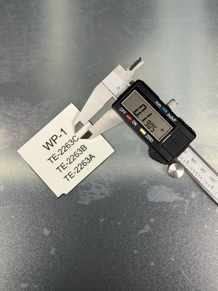 Digital calipers measuring the thickness of a white phenolic tag engraved WP-1